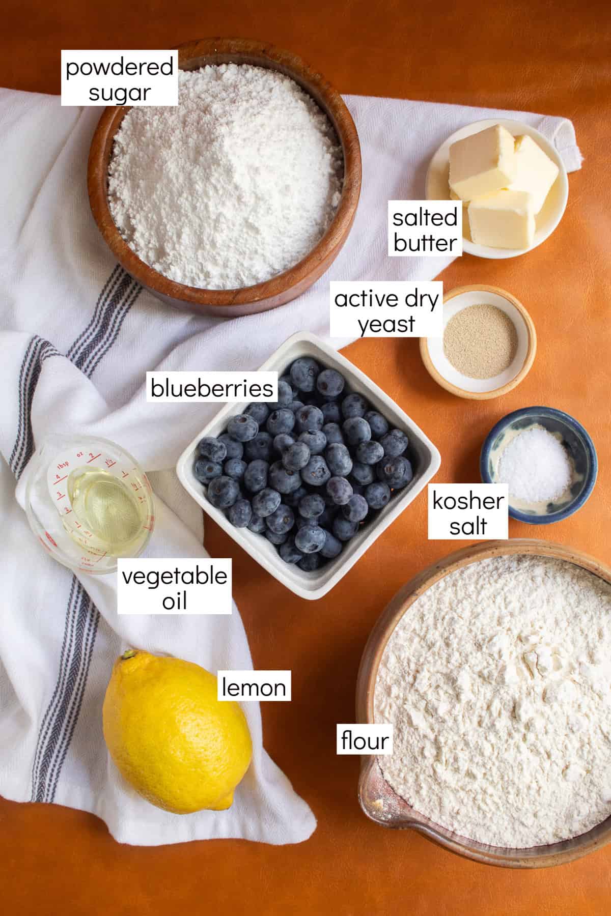 Ingredients for the recipe are displayed and labeled with text boxes including powdered sugar, salted butter, active dry yeast, blueberries, kosher salt, vegetable oil, lemon, and flour.