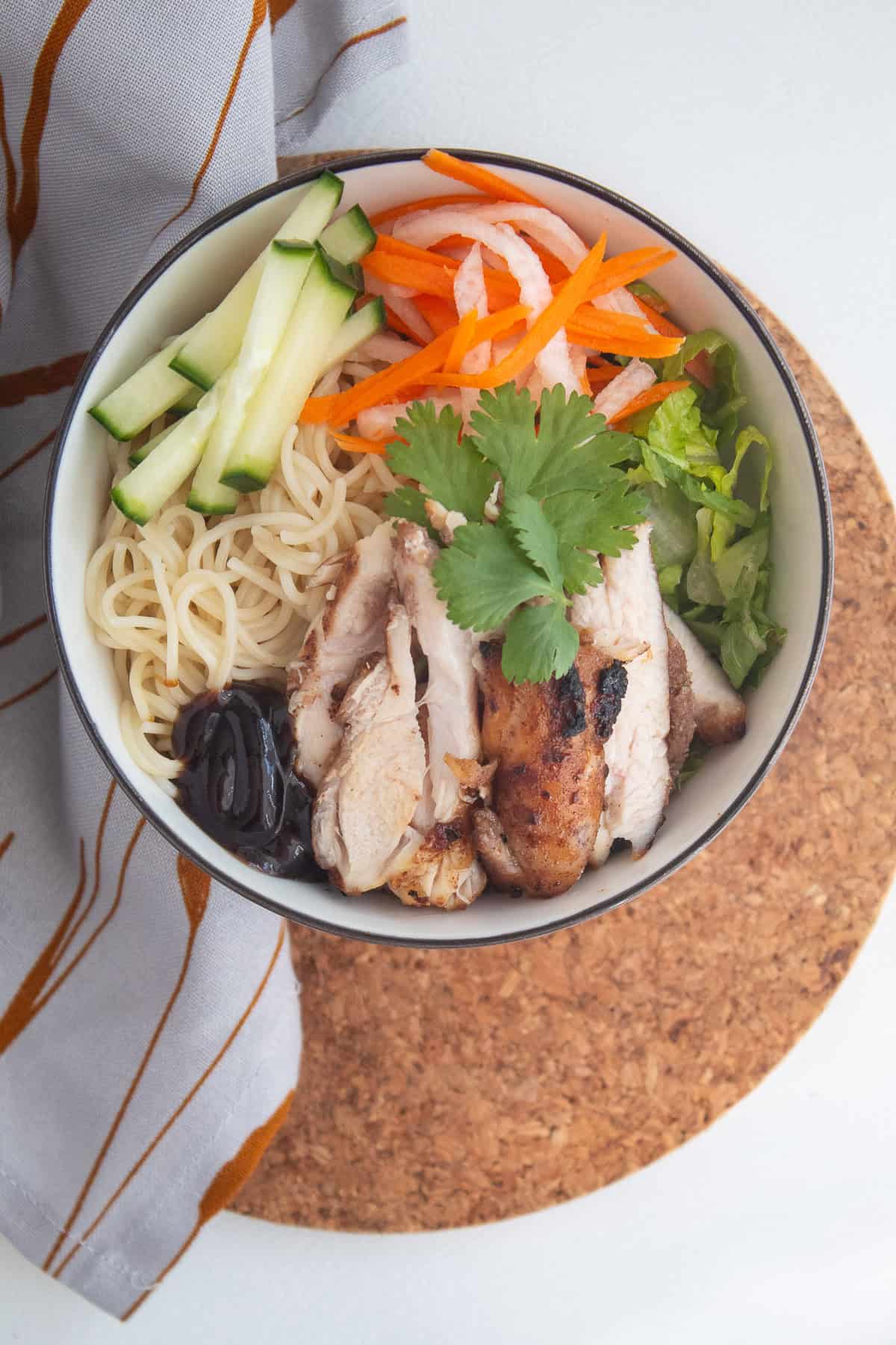 A noodle bowl is topped with shredded lettuce, cilantro, hoisin sauce, cucumber sticks, pickled vegetables, and 5 spice chicken.