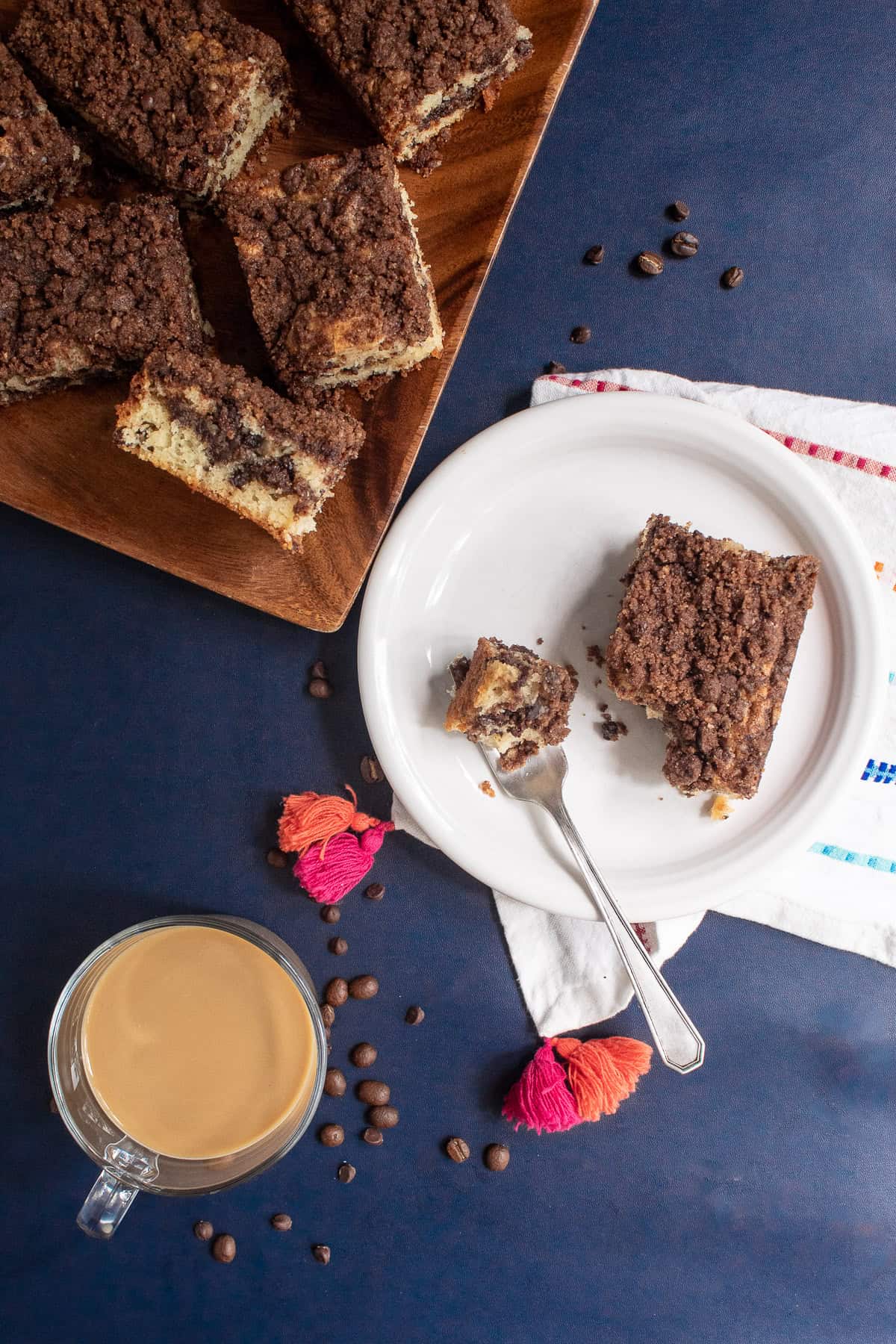 A serving of coffee cake on a white plate is next to a platter of slices and a cup of coffee.