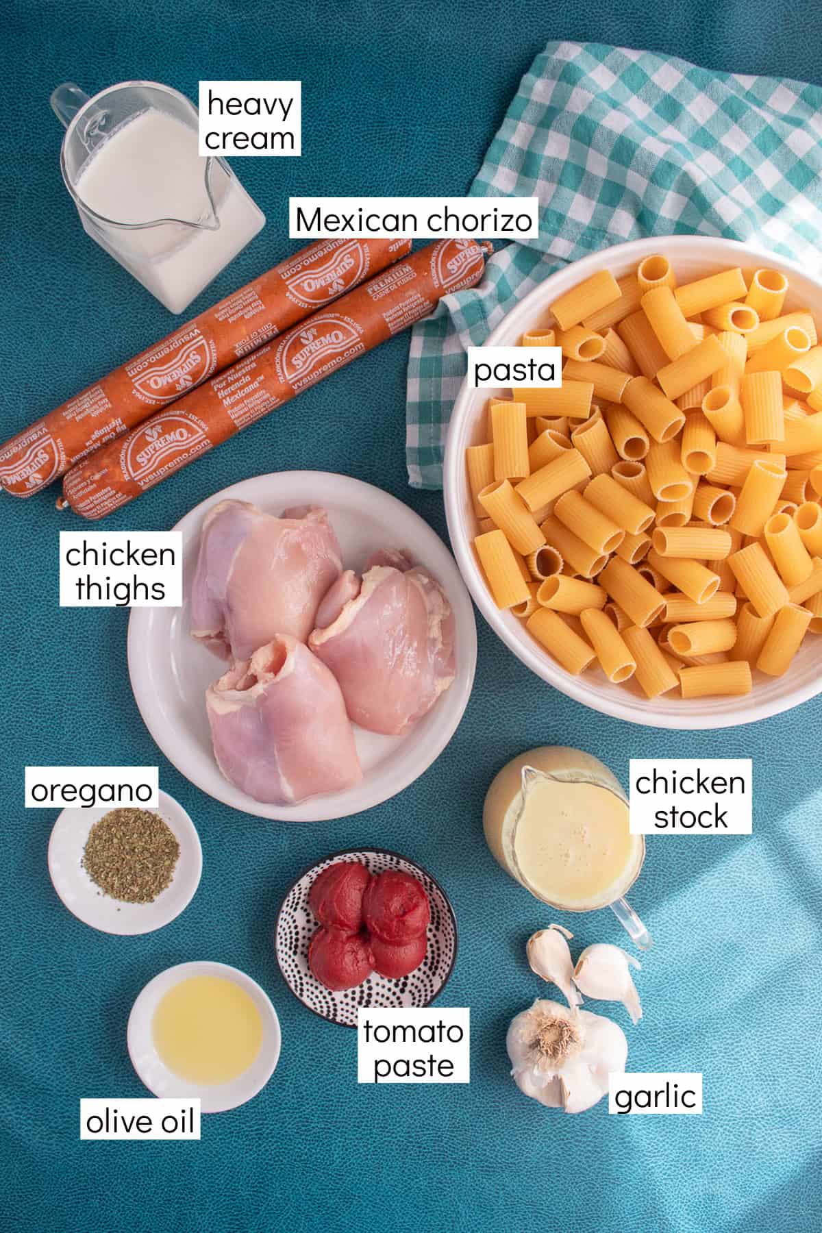Ingredients for the recipe are displayed and labeled with white text boxes including the words heavy cream, Mexican chorizo, pasta, chicken thighs, oregano, chicken stock, olive oil, tomato paste, and garlic.
