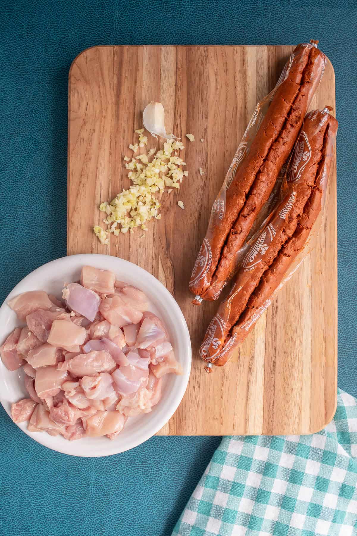 Chicken thighs cut into bite-sized pieces, minced garlic, and chorizo removed from the casing are displayed on a wooden cutting board.