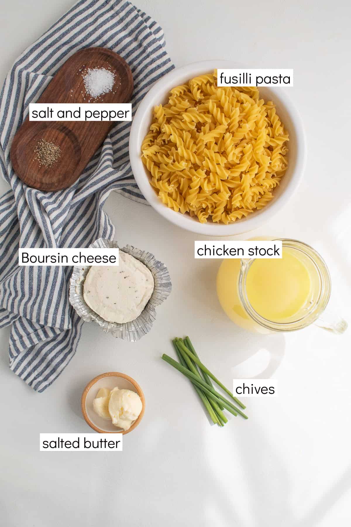 Ingredients for the recipe are displayed and labeled with white text boxes including the words salt and pepper, fusilli pasta, chicken stock, Boursin cheese, salted butter, and chives.