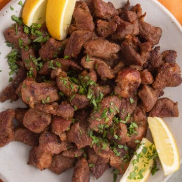 Pieces of lamb are served on a platter and garnished with herbs and lemon wedges.