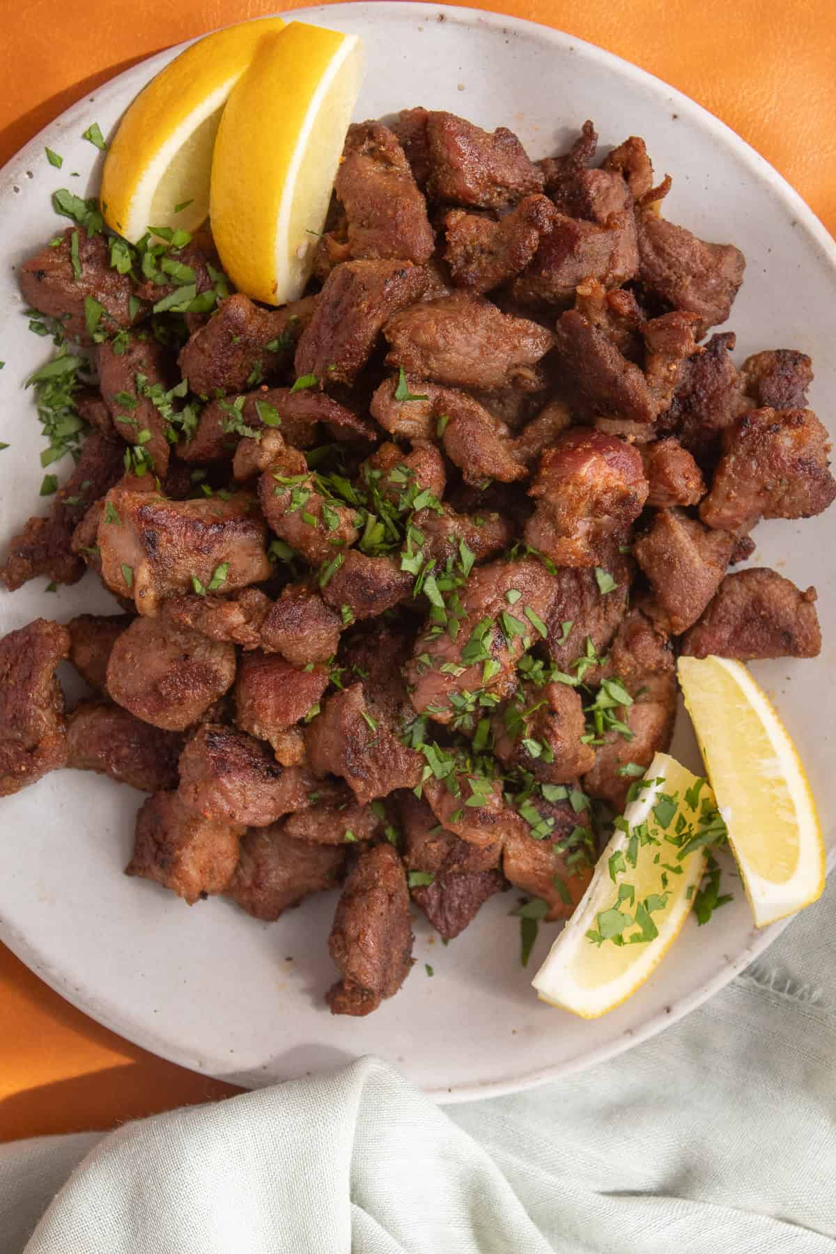 Detail image of pieces of spiced lamb served on a platter and garnished with herbs and lemon wedges.