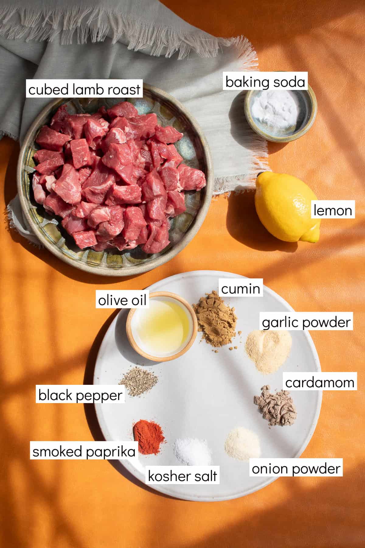 Ingredients for the recipe are displayed and labeled with white text boxes including the words cubed lamb roast, baking soda, lemon, cumin, garlic powder, cardamom, onion powder, kosher salt, smoked paprika, black pepper, and olive oil.