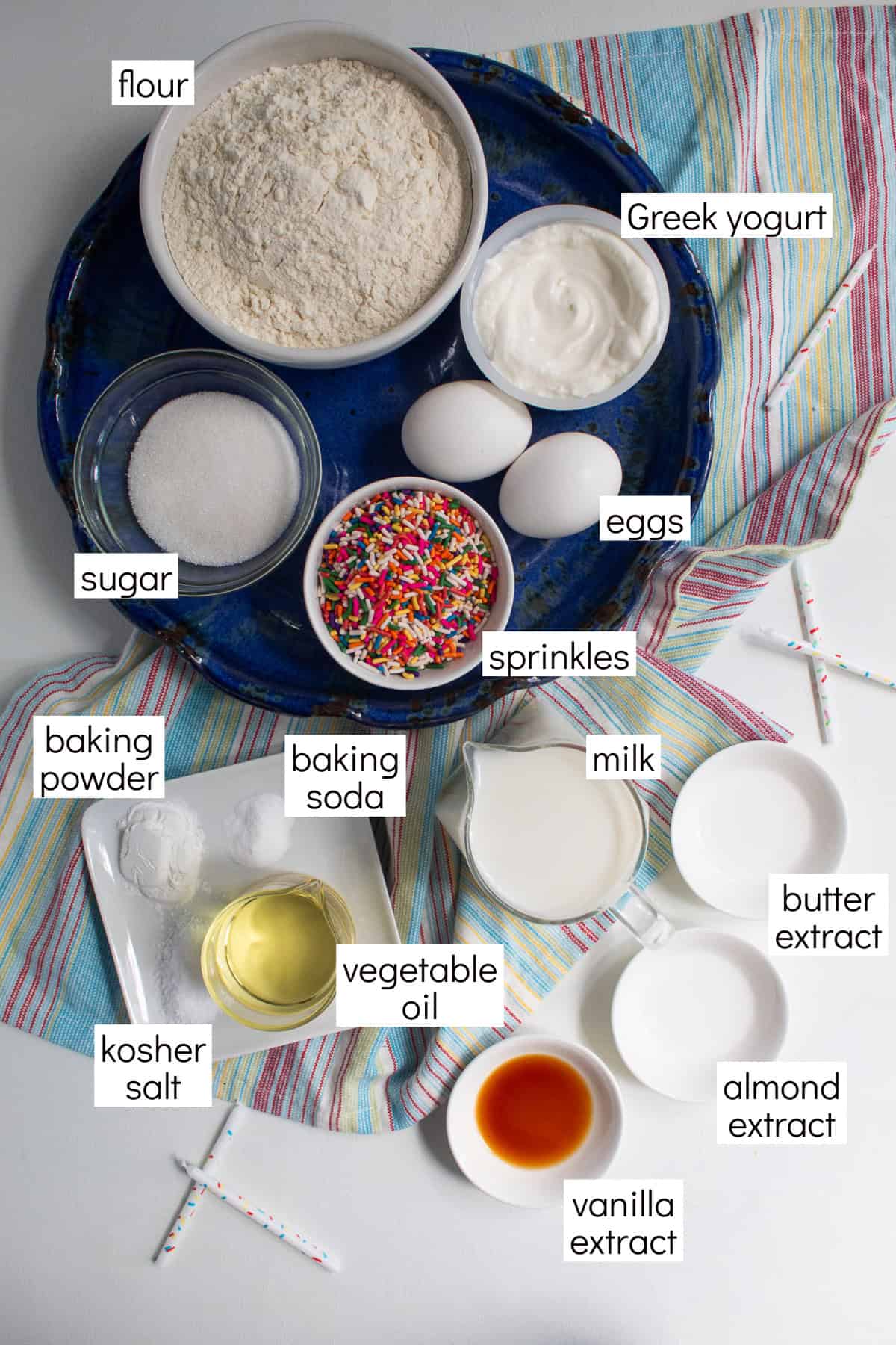 Ingredients for the recipe are displayed and labeled with white text boxes including the words flour, Greek yogurt, sugar, eggs, sprinkles, baking powder, baking soda, milk. kosher salt, vegetable oil, butter extract, almond extract, and vanilla extract.