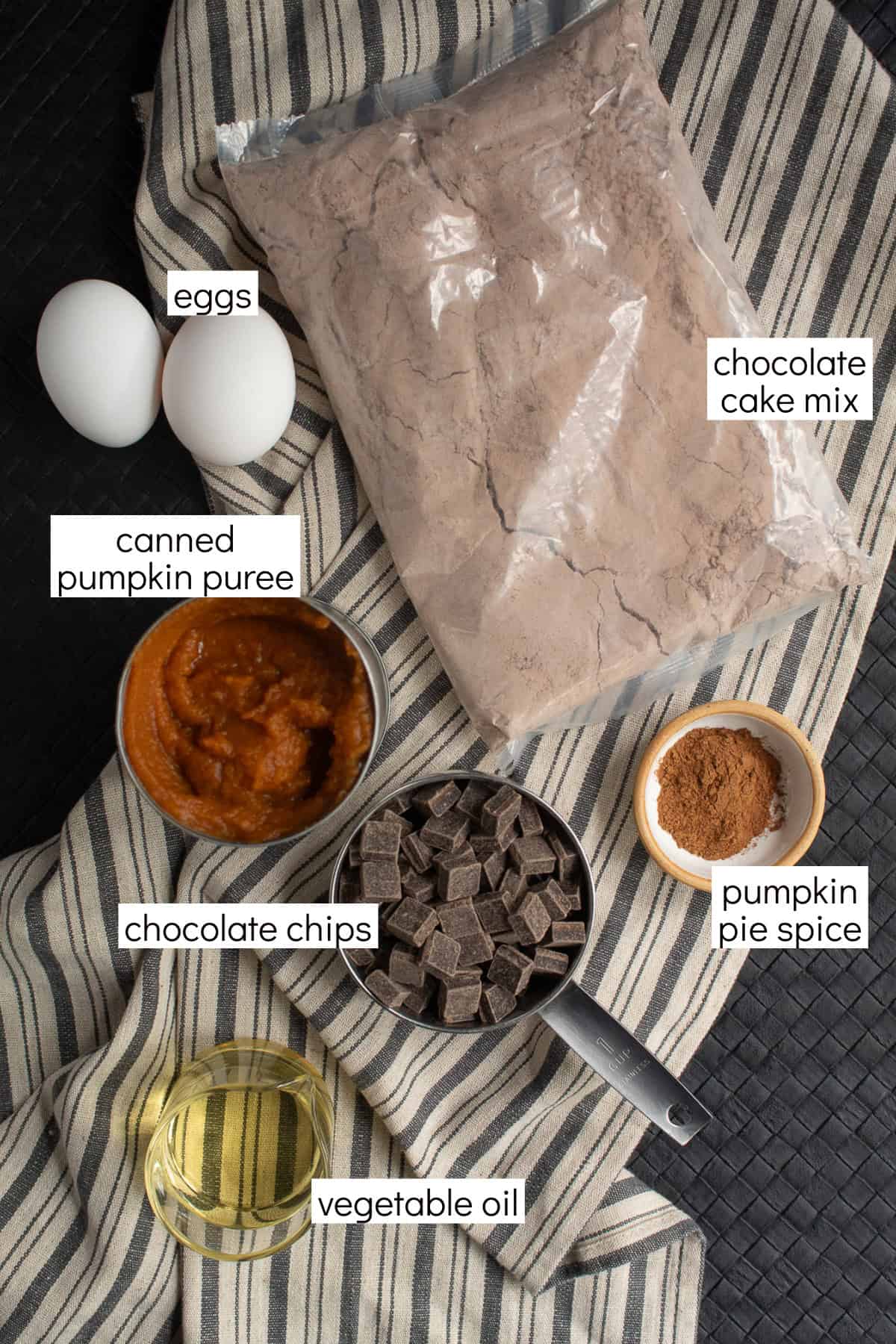Ingredients for the recipe are displayed and labeled with white text boxes including the words eggs, chocolate cake mix, canned pumpkin puree, chocolate chips, pumpkin pie spice, and vegetable oil.