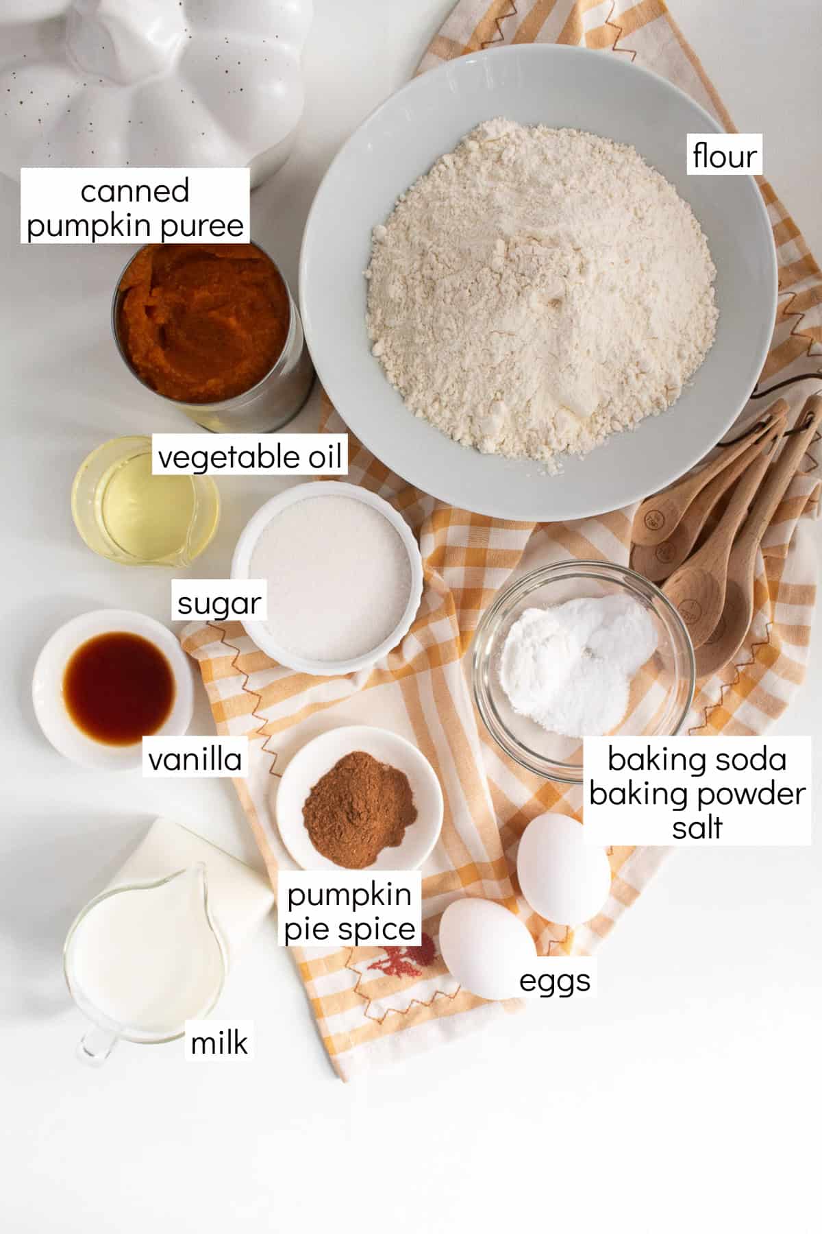 Ingredients for the pancakes are displayed and labeled with white text boxes including the words canned pumpkin puree, flour, vegetable oil, sugar, vanilla, pumpkin pie spice, milk, eggs, and baking soda, baking powder, and salt.