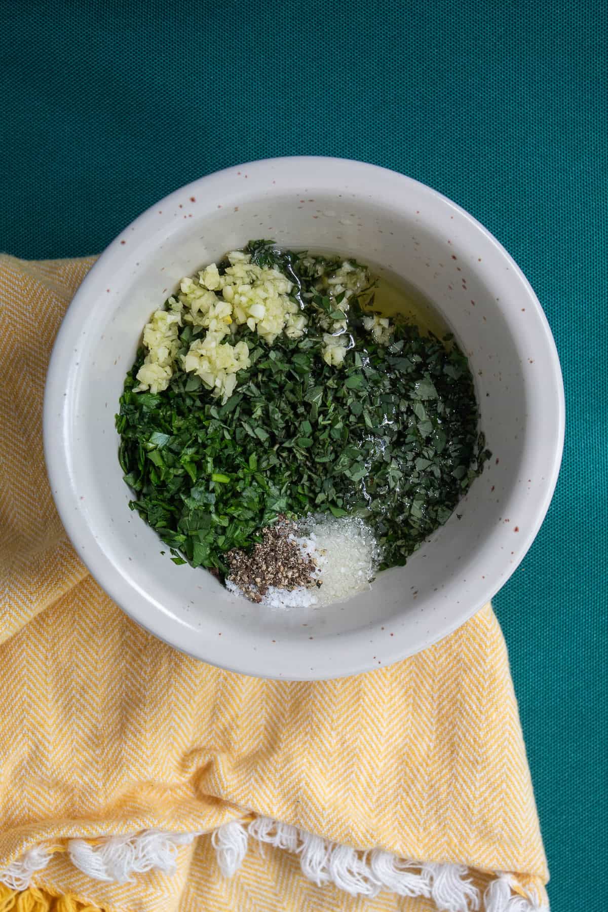 Chimichurri ingredients, including garlic, parsley, oregano, salt, pepper, red vine vinegar, and olive oil, are piled into a small bowl.