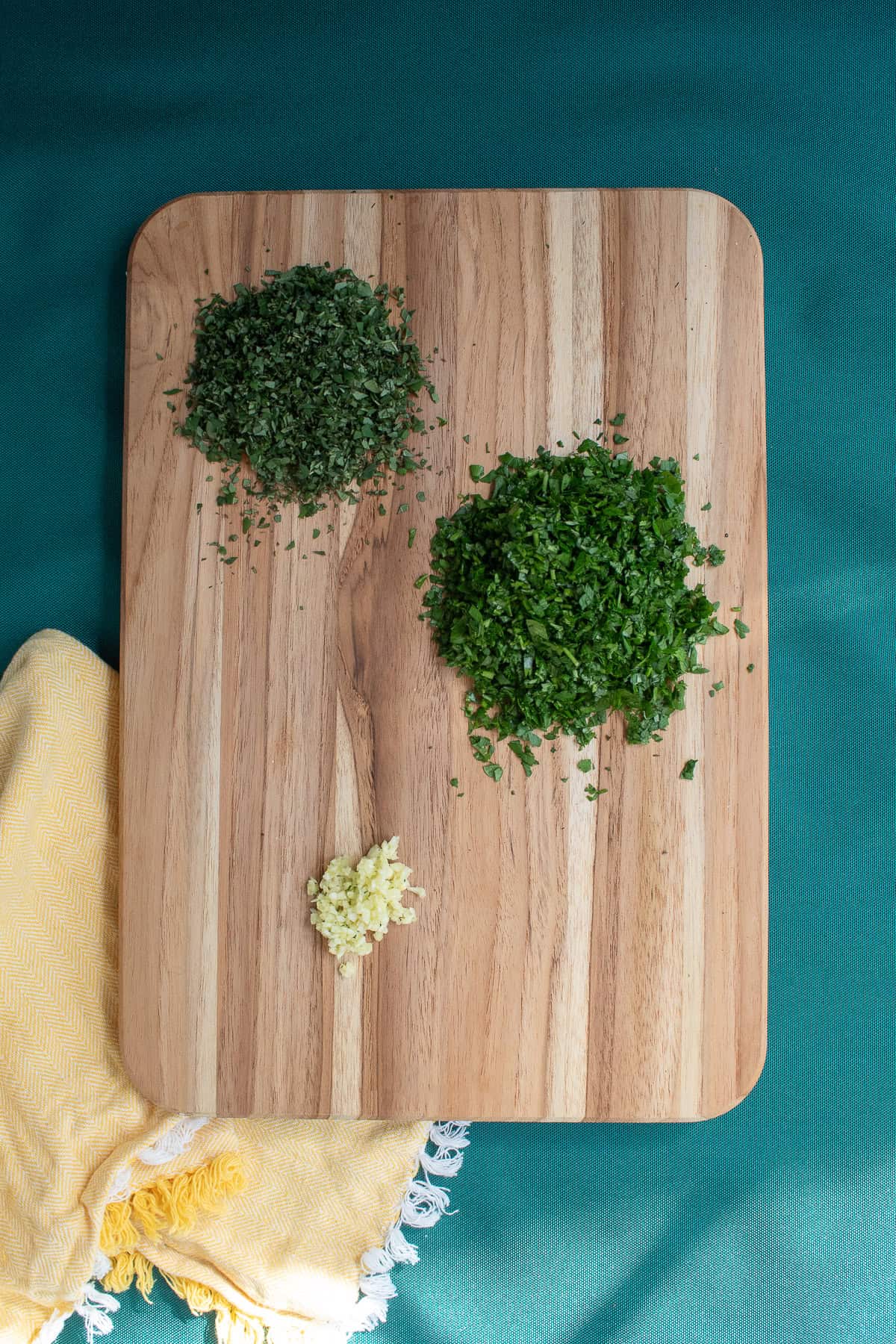 Piles of finely minced oregano leaves, parsley, and garlic sit on a wooden cutting board.
