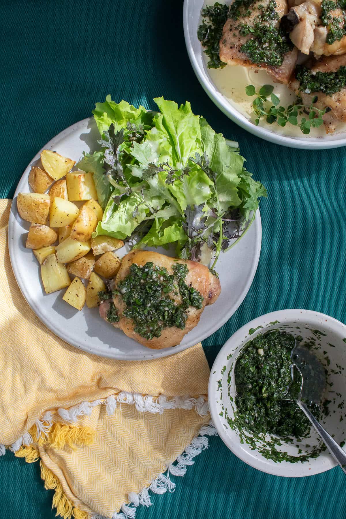 A dinner plate features chimichurri sauce on a chicken thigh, roasted potatoes, and a green salad.