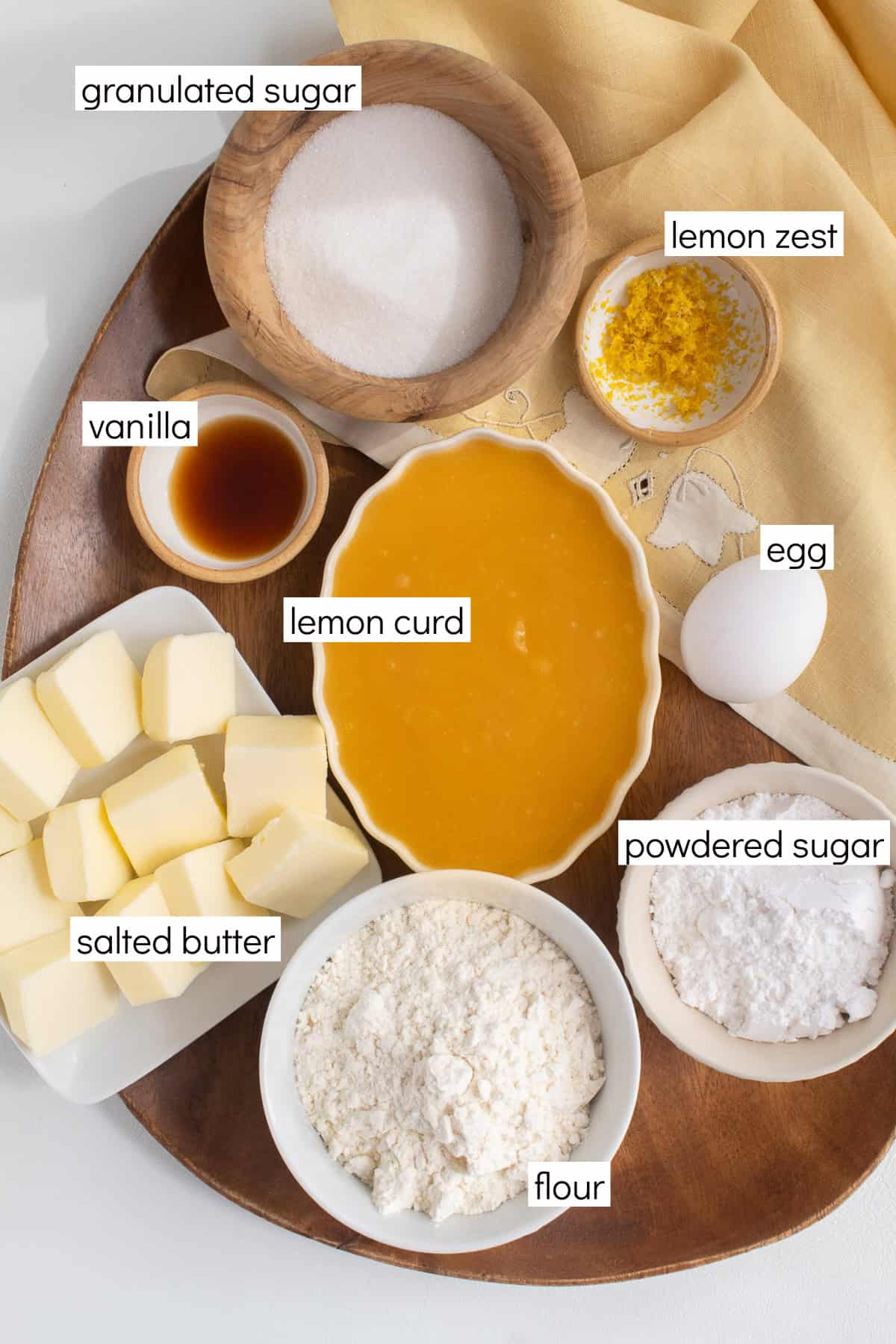 Ingredients for the recipe are displayed and labeled with white text boxes including the words granulated sugar, lemon zest, vanilla, lemon curd, egg, salted butter, powdered sugar, and flour.