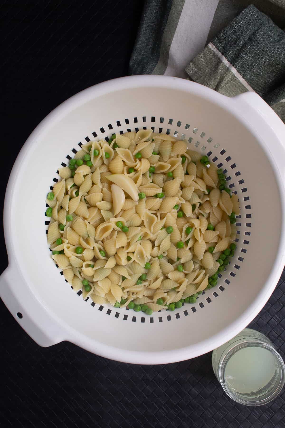 The pasta, peas, and garlic are strained in a plastic colander, while some pasta water is reserved.