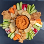 A platter of vegetables and crackers surrounds the vibrant muhammara swirled in a silver bowl.