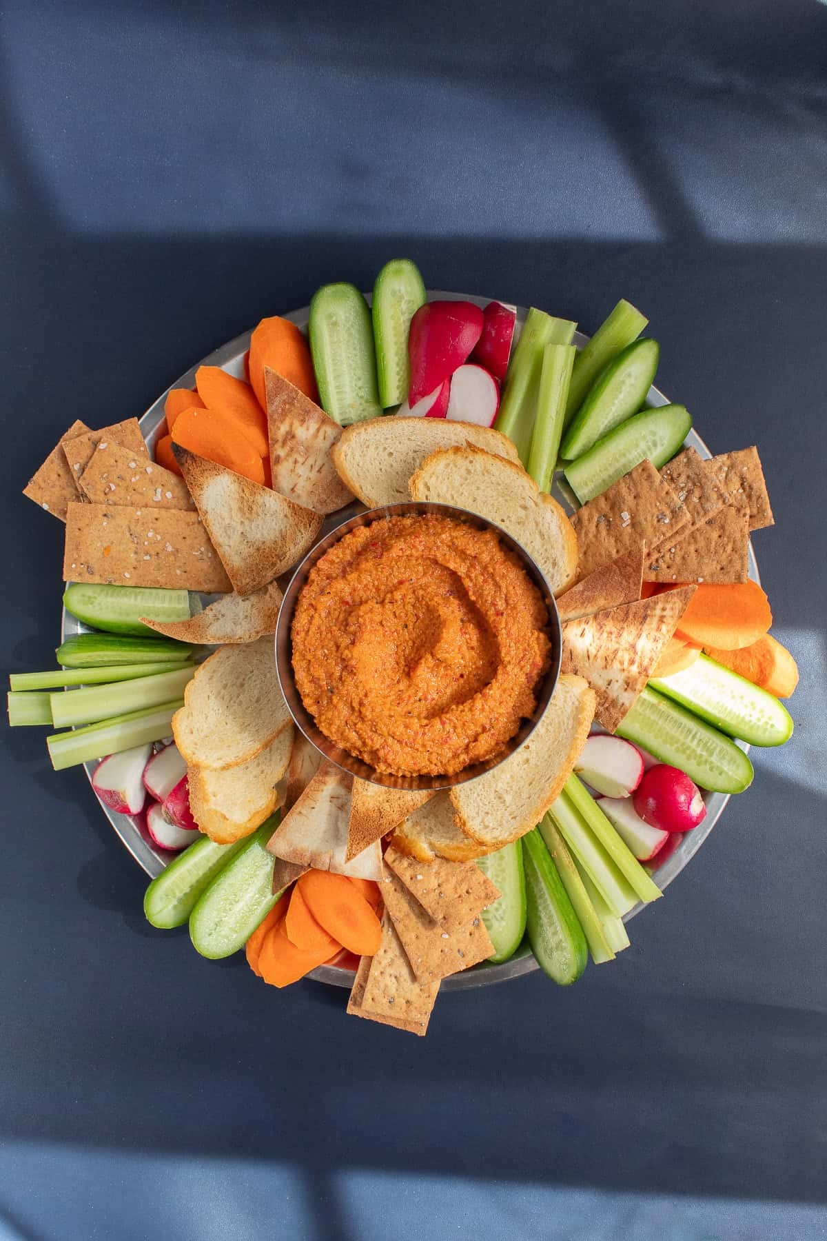 A platter of vegetables and crackers surrounds the vibrant muhammara swirled in a silver bowl.