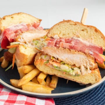 The sandwich is cut in half, showing the layers of sauce, slaw, chicken breast, and tomato on the brioche bun.