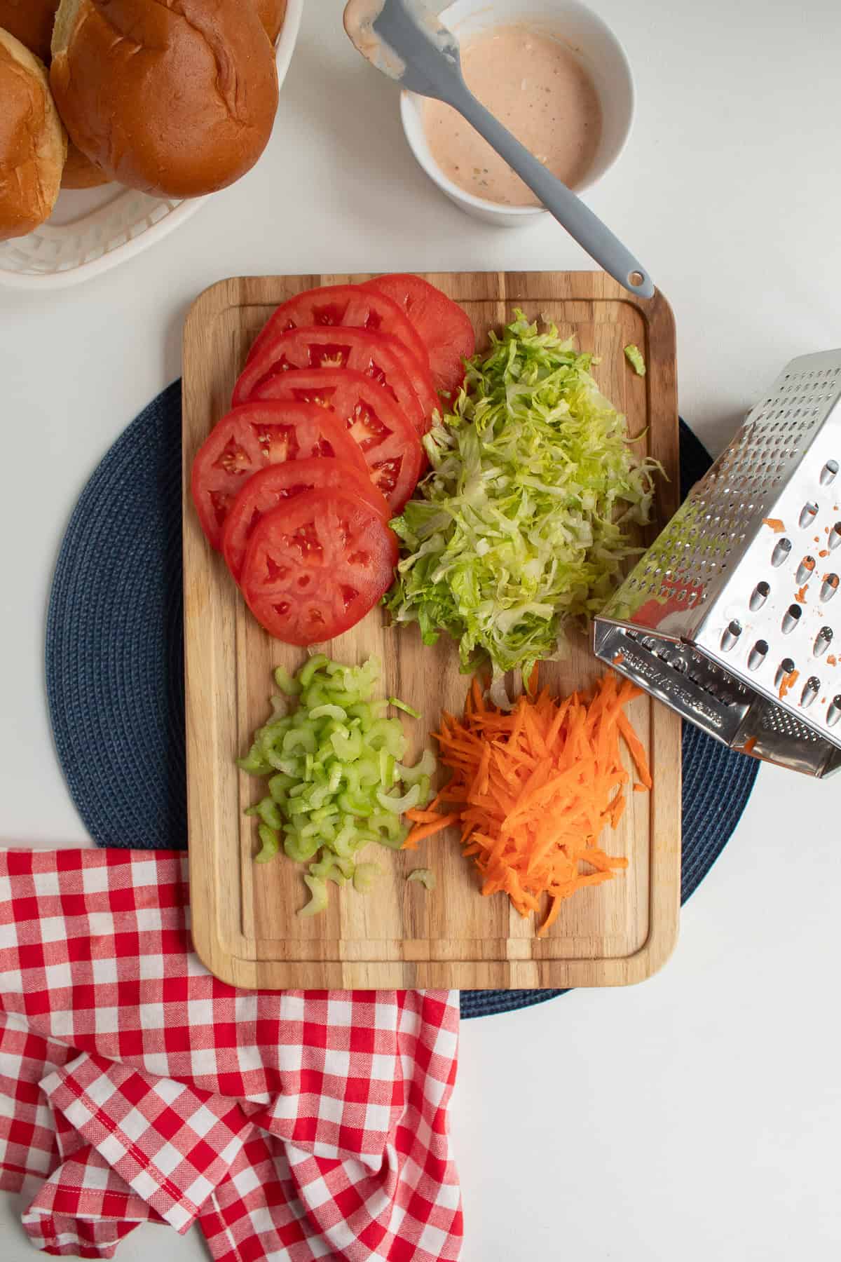 Tomatoes, lettuce, celery, and carrots are prepared on a wooden cutting board for sandwich toppings
