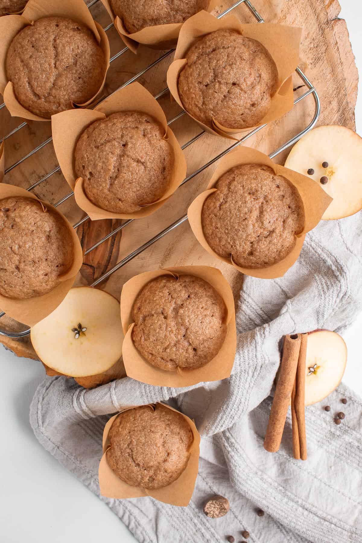 Baked muffins cool on a wire rack and are surrounded by apple slices, cinnamon, allspice, and nutmeg.