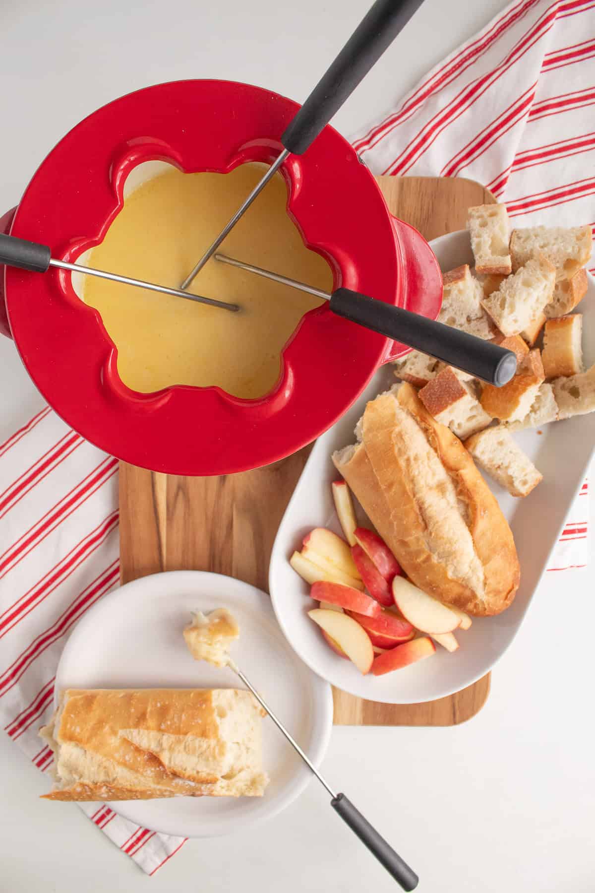 A red fondue pot holds three forks and is placed near a platter of bread and apple slices.
