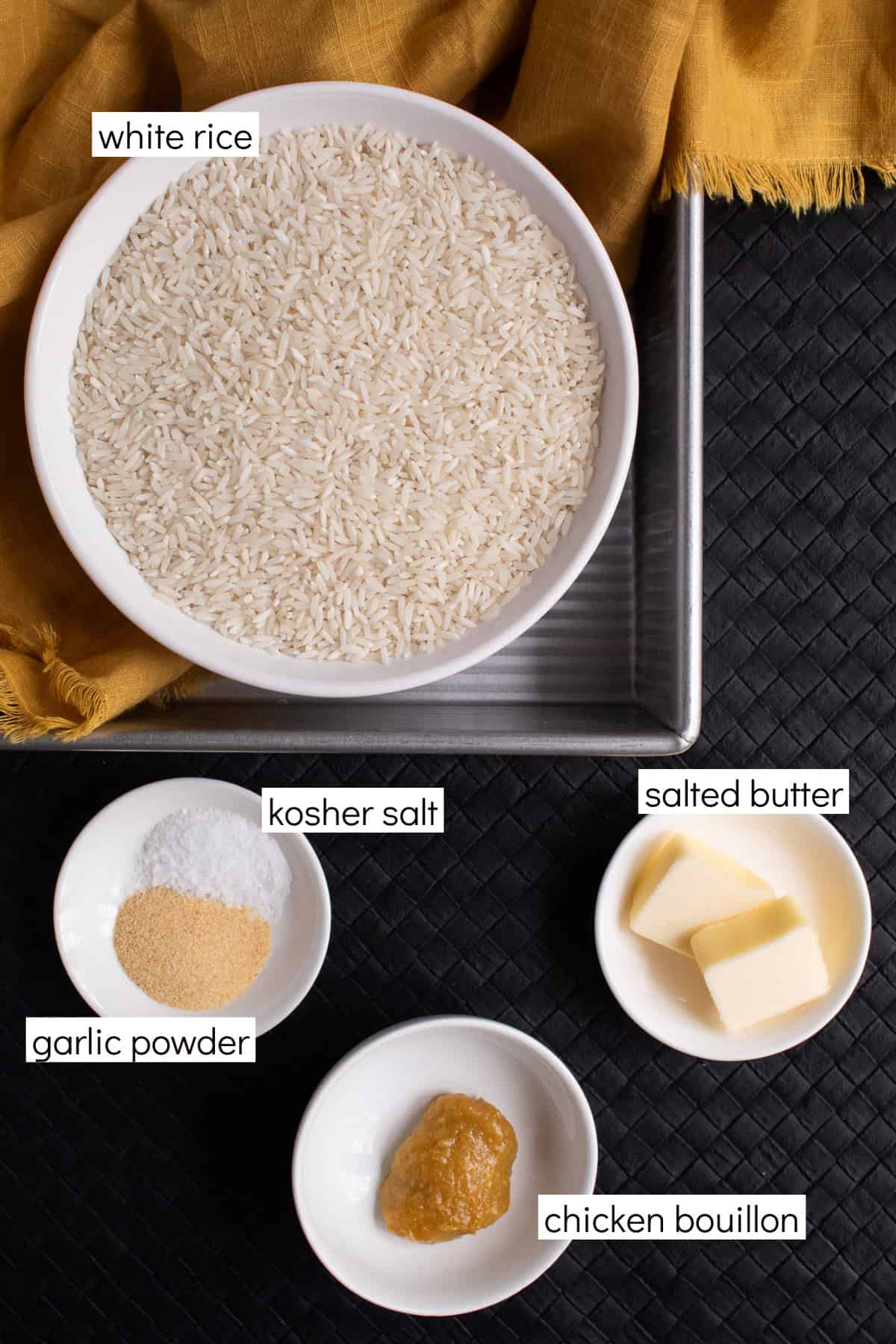 Ingredients for the recipe are displayed and labeled with white text boxes including the words white rice, kosher salt, salted butter, garlic powder, and chicken bouillon.