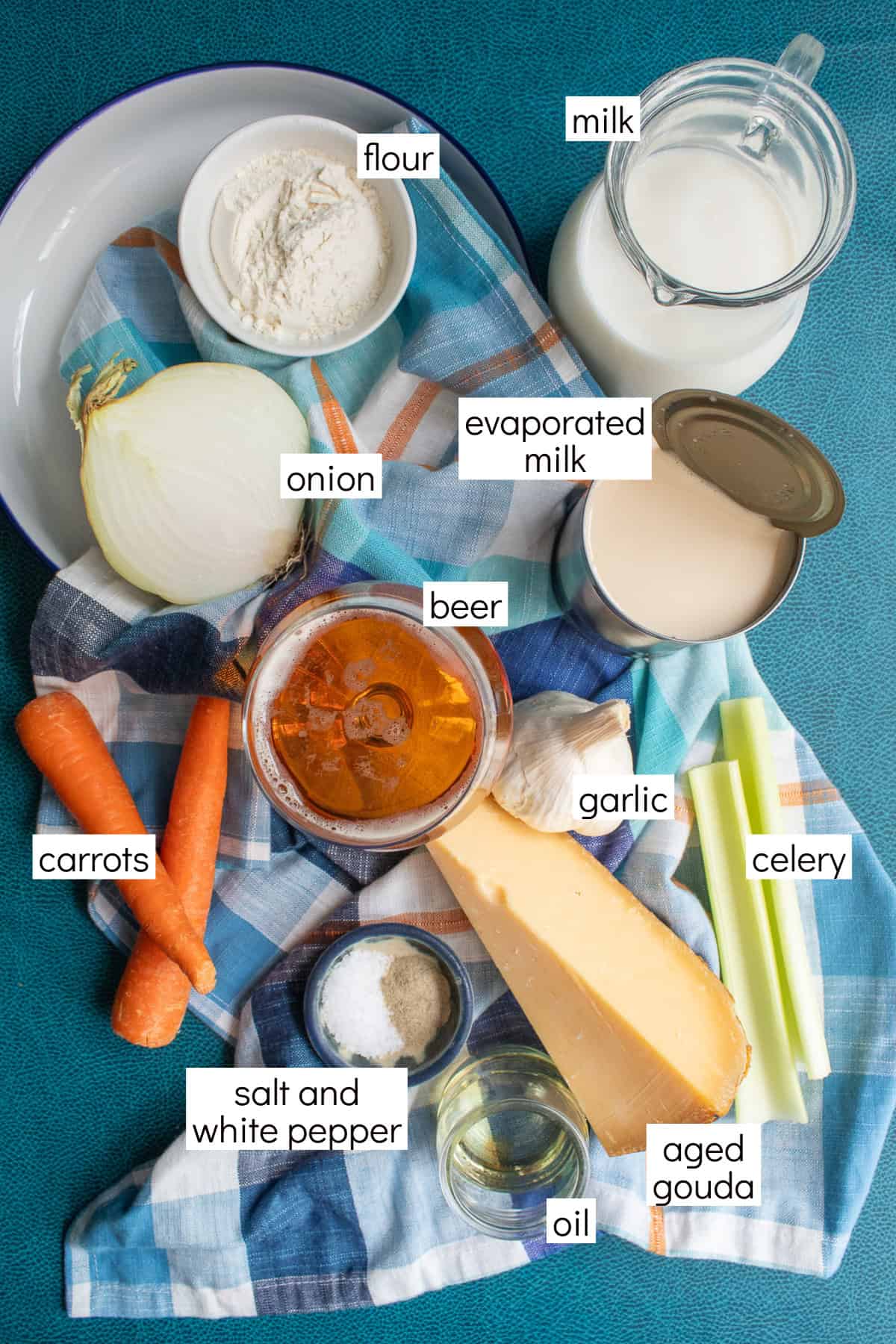Ingredients for the recipe are displayed and labeled with white text boxes including the words flour, milk, onion, evaporated milk, beer, carrots, garlic, celery, salt and white pepper, oil, and aged gouda.