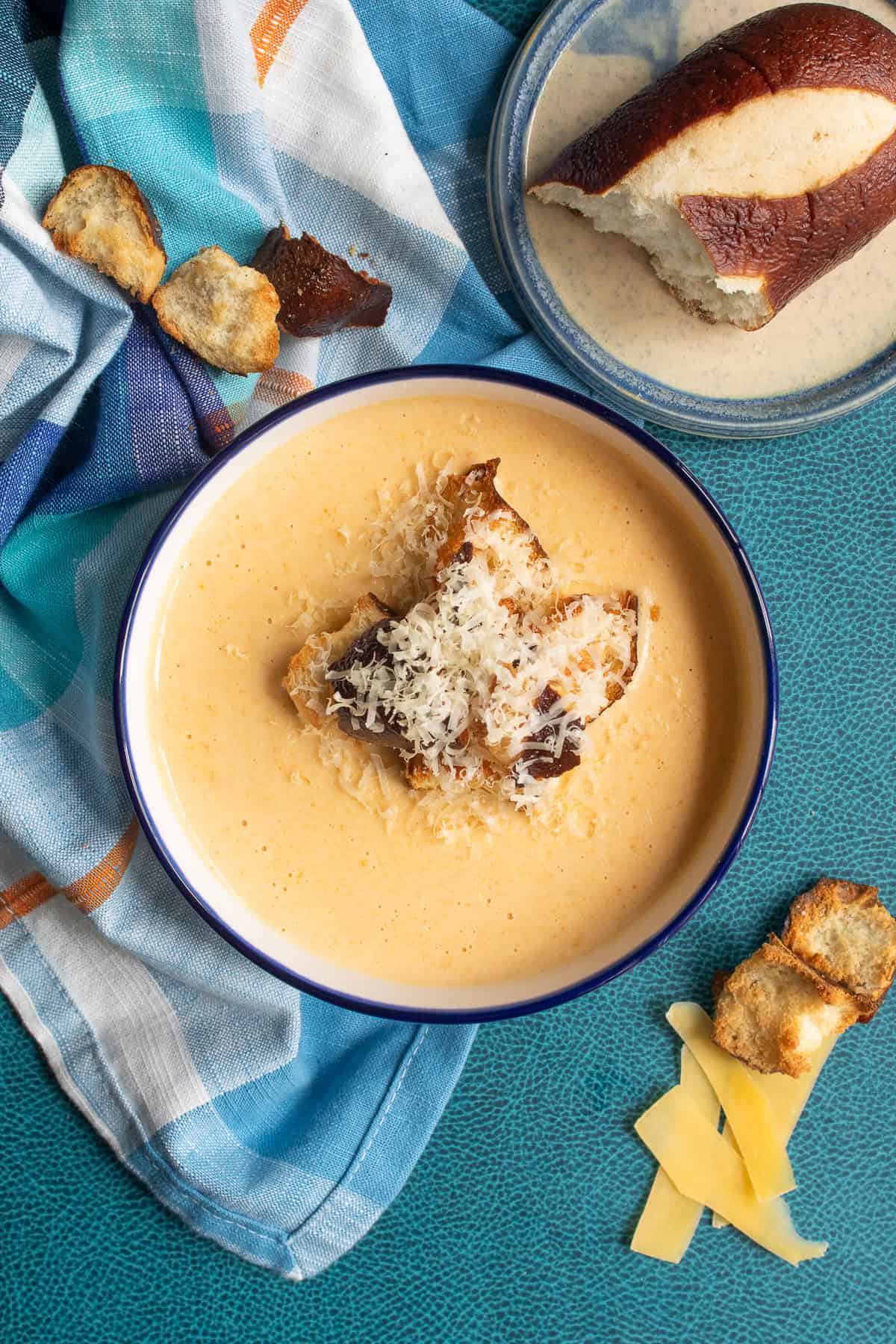 The creamy soup is displayed on a blue surface and is topped with pretzel bread croutons and additional shredded gouda cheese.