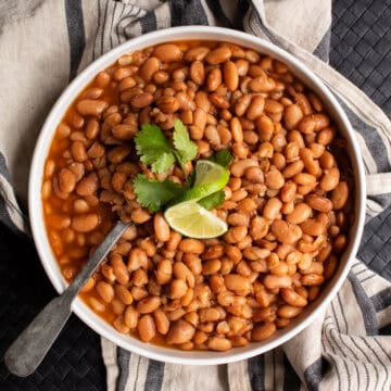 A white bowl is filled with cooked pinto beans and garnished with lime and cilantro.