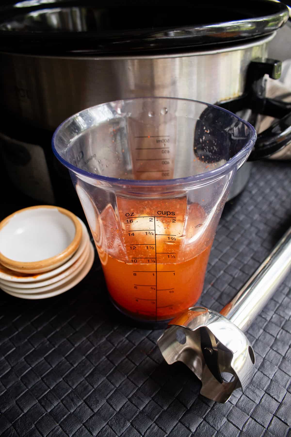 An orange liquid in a blender cup sits on a black surface next to an immersion blender.