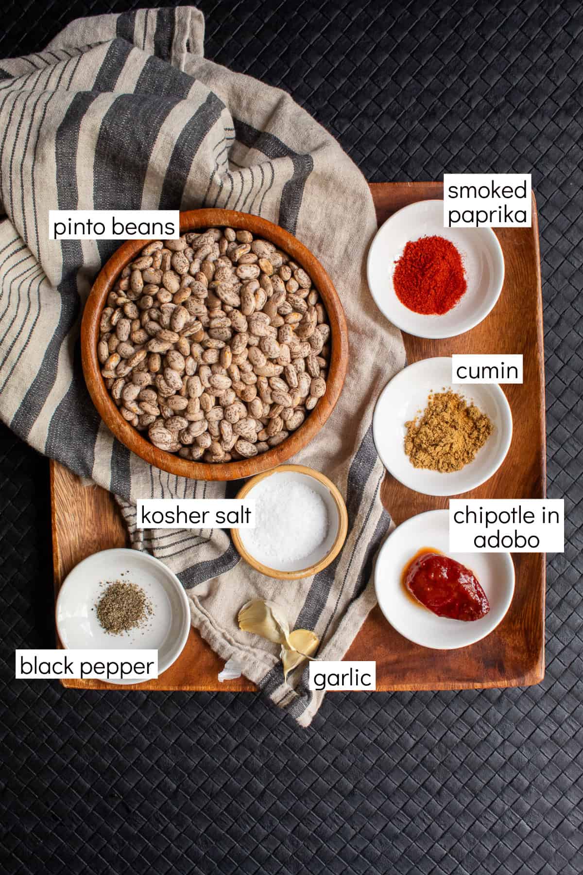 Ingredients for the recipe are displayed and labeled with white text boxes including the words pinto beans, smoked paprika, cumin, chipotle in adobo, kosher salt, black pepper, and garlic.