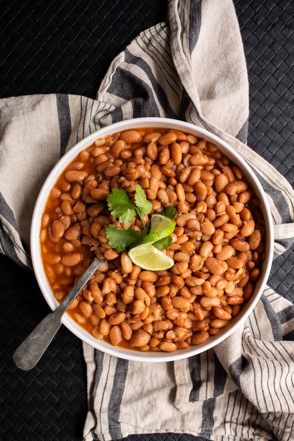 A white bowl piled with cooked pinto beans is garnished with cilantro and lime.