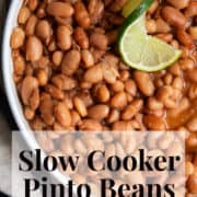 A white platter is filled with cooked pinto beans and garnished with lime. A white text box at the bottom of the image contains the words, "slow cooker pinto beans" and "plan. eat. post. repeat."