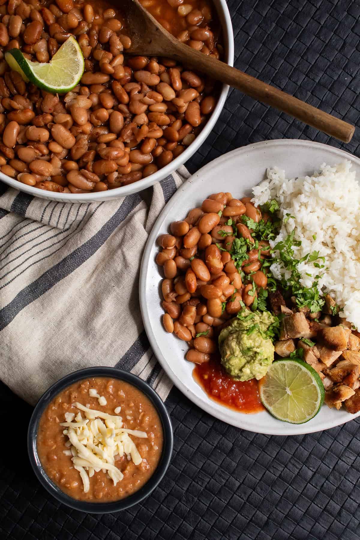 A platter of cooked beans sits next to a serving of beans, rice, and pork as well as a bowl of refried beans.