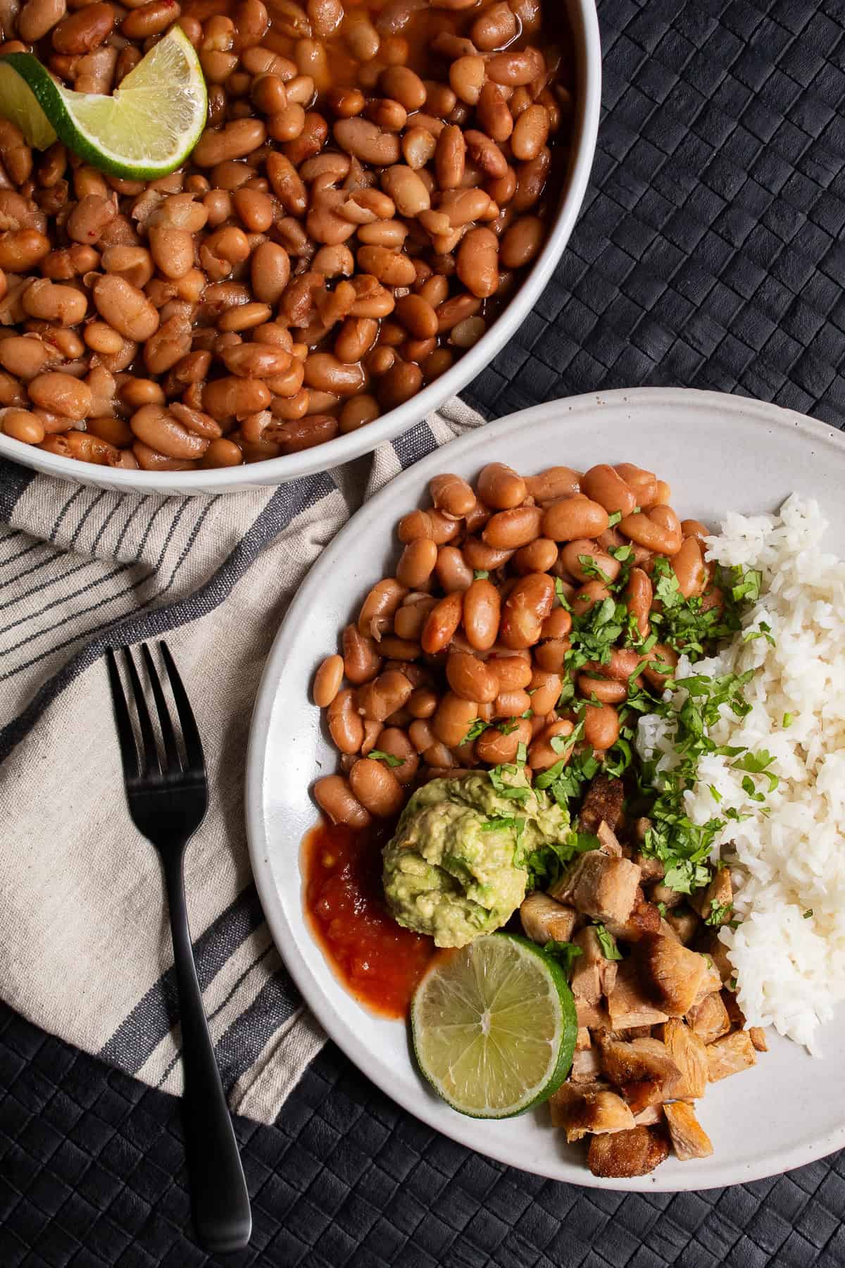 A plate of pinto beans, rice, carnitas, guacamole, and salsa is garnished with cilantro and lime.