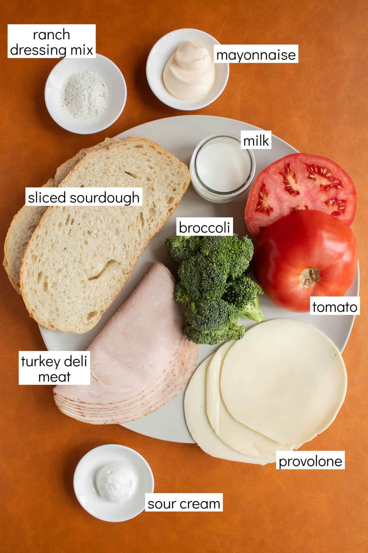 Ingredients for the recipe are displayed and labeled with white text boxes including the words ranch dressing mix, mayonnaise, milk, sliced sourdough, broccoli, tomato, turkey deli meat, provolone, and sour cream.