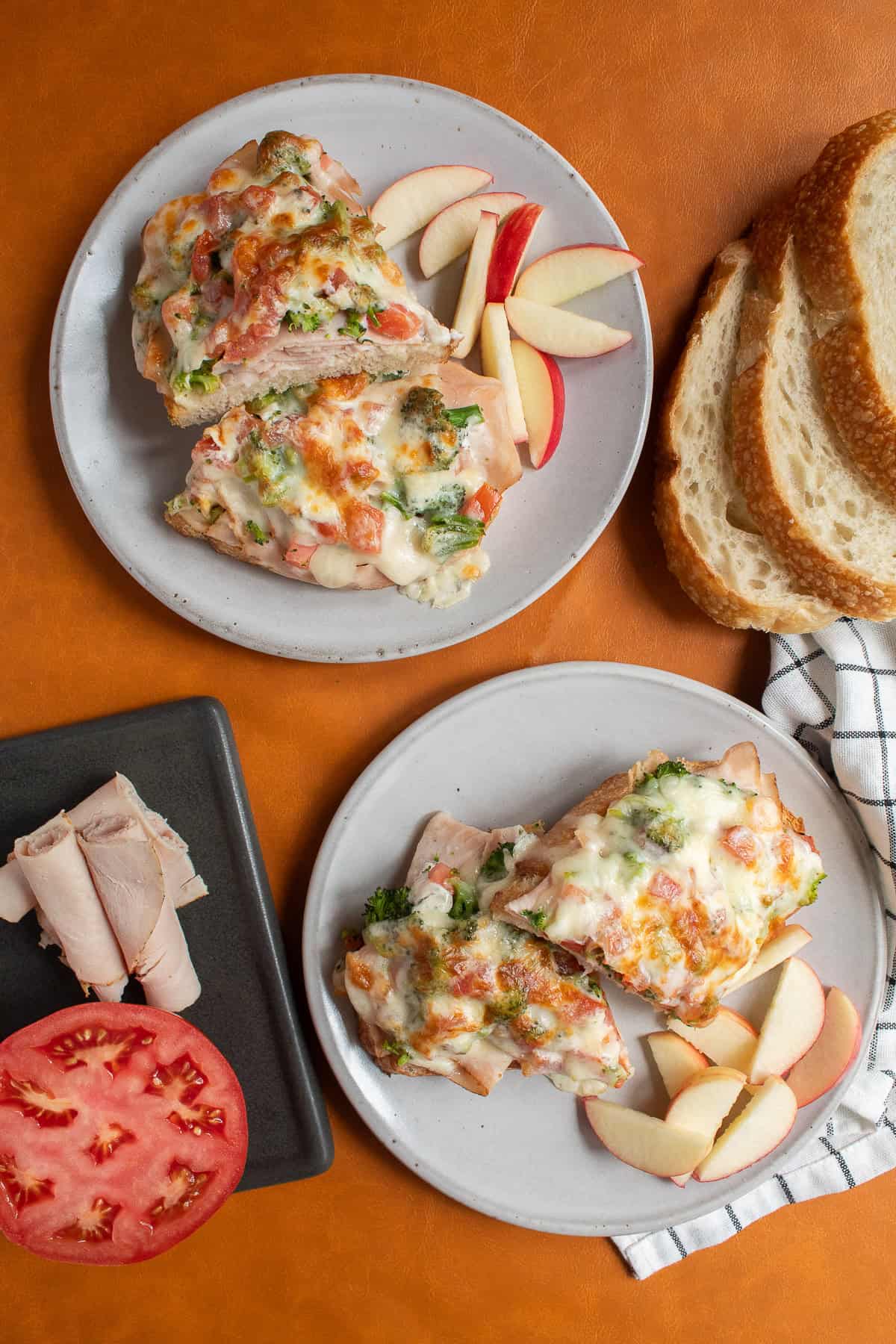 Two plates are stacked with halved turkey melts and apple slices.