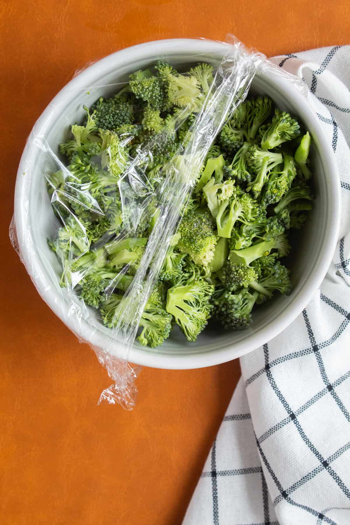 Chopped broccoli is in a white bowl partially covered with plastic wrap.