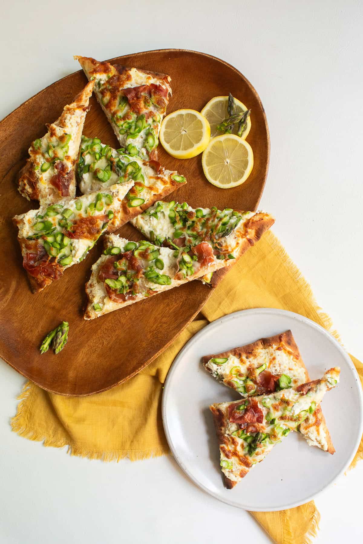 Pizza slices are piled on a wooden platter alongside lemon slices and asparagus tips.