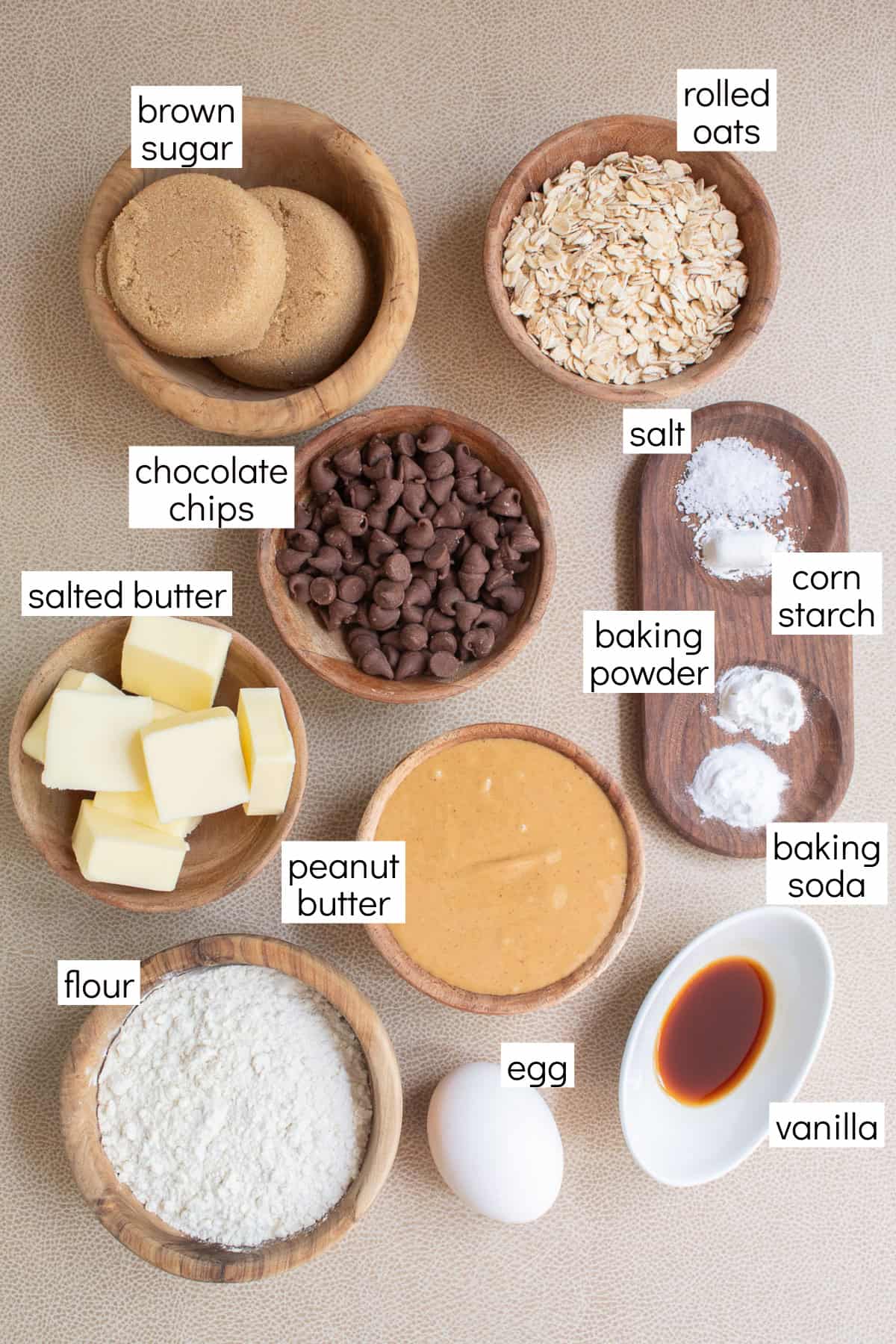 Ingredients for the recipe are displayed and labeled with white text boxes including the words brown sugar, rolled oats, chocolate chips, salt, salted butter, corn starch baking powder, peanut butter, baking soda, flour, egg, and vanilla.