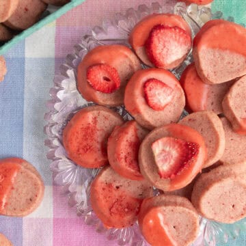A glass platter of strawberry shortbread cookies is decorated with dried strawberries, strawberry glaze, and crushed strawberry powder.