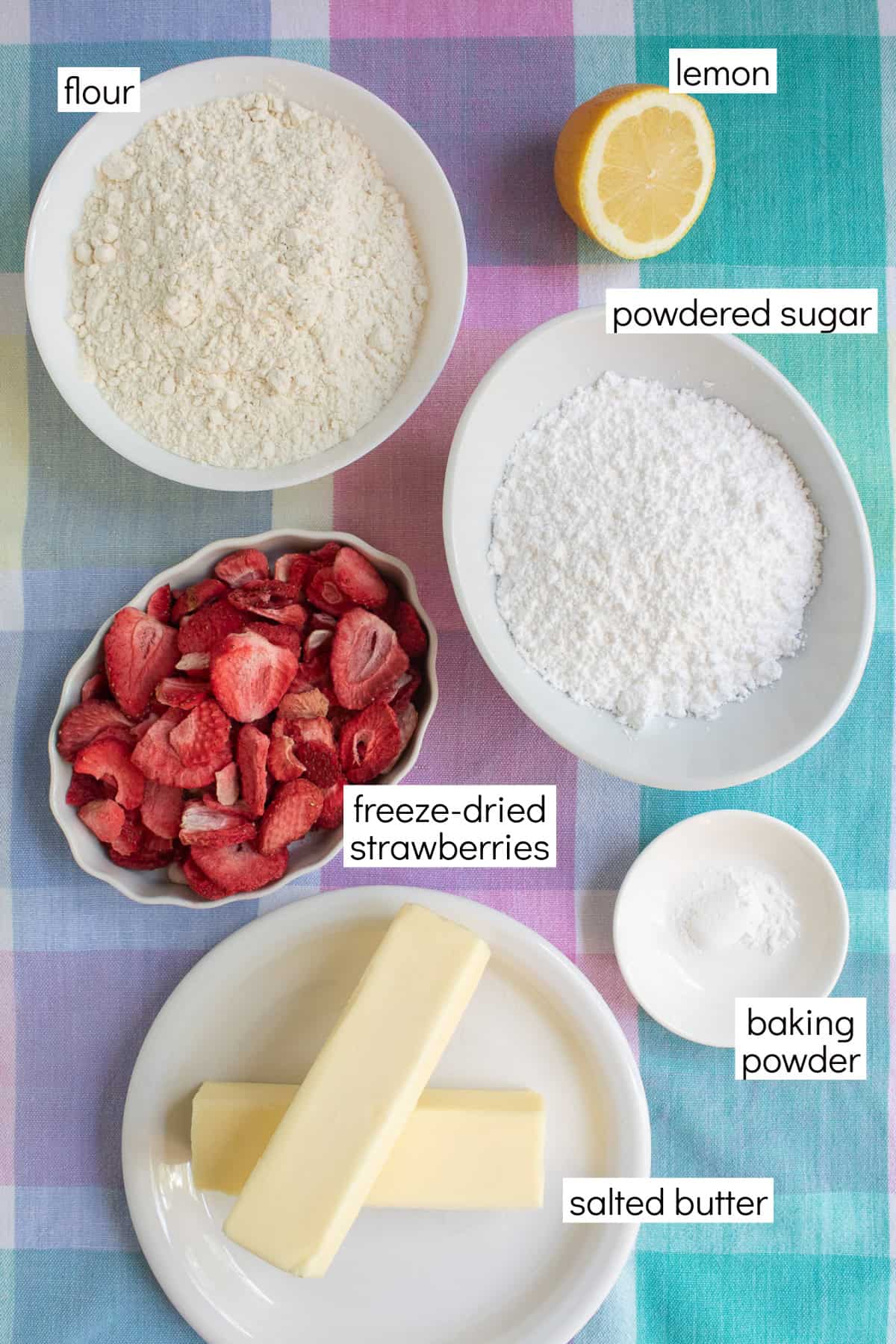 Ingredients for the recipe are displayed and labeled with white text boxes including the words flour, lemon, powdered sugar, freeze-dried strawberries, baking powder, and salted butter.