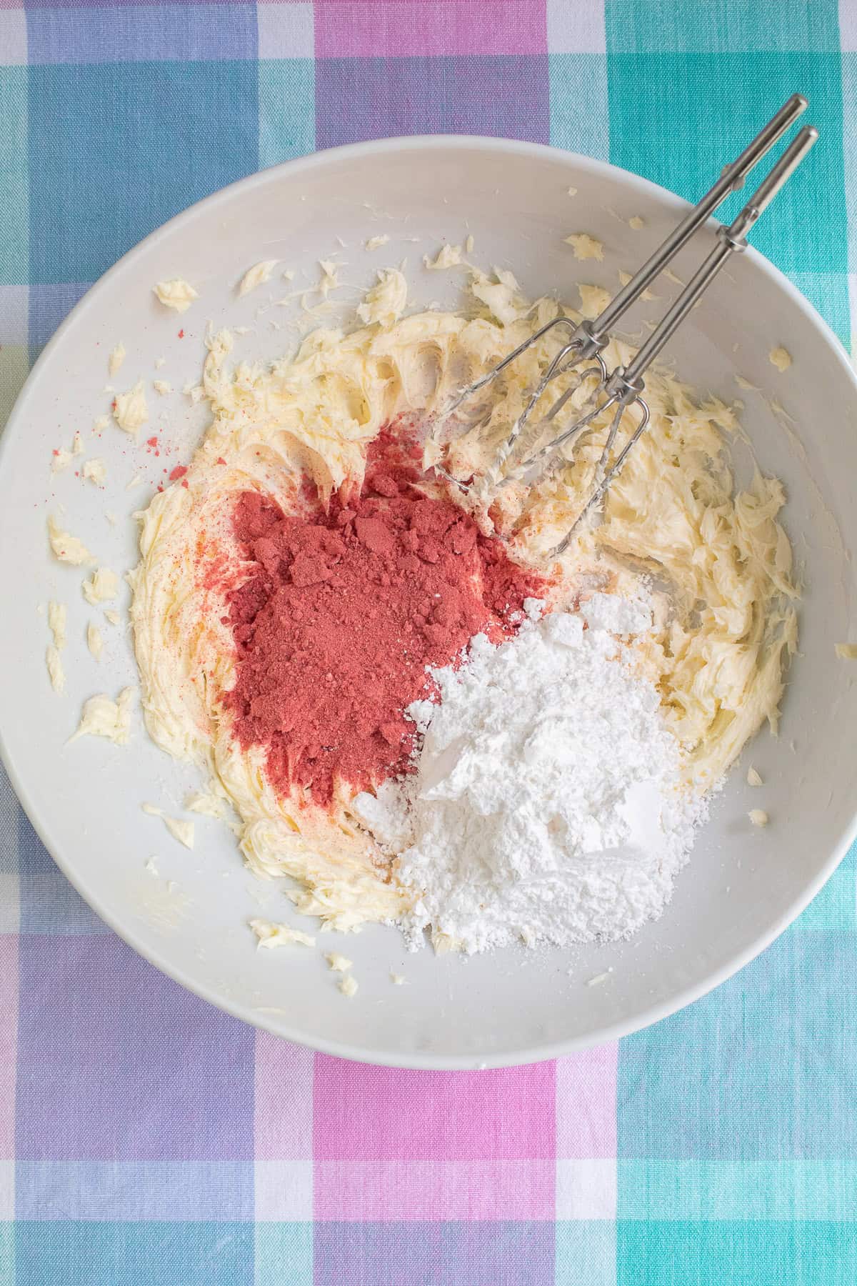 Dried strawberry powder and powdered sugar are added to creamed butter in a large white mixing bowl.