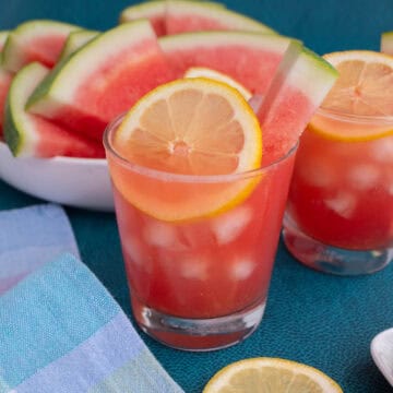 A rosy hued glass of watermelon lemonade is garnished with a lemon slice and watermelon spear.