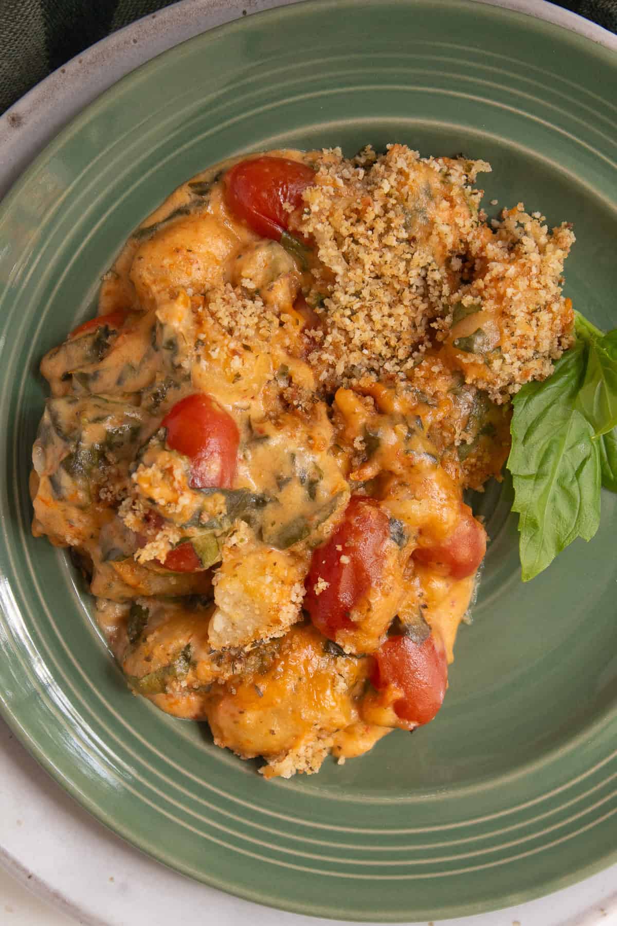 A detail image of a green plate loaded with baked gnocchi with breadcrumbs, grape tomatoes, basil, and spinach visible in the sun dried tomato sauce.