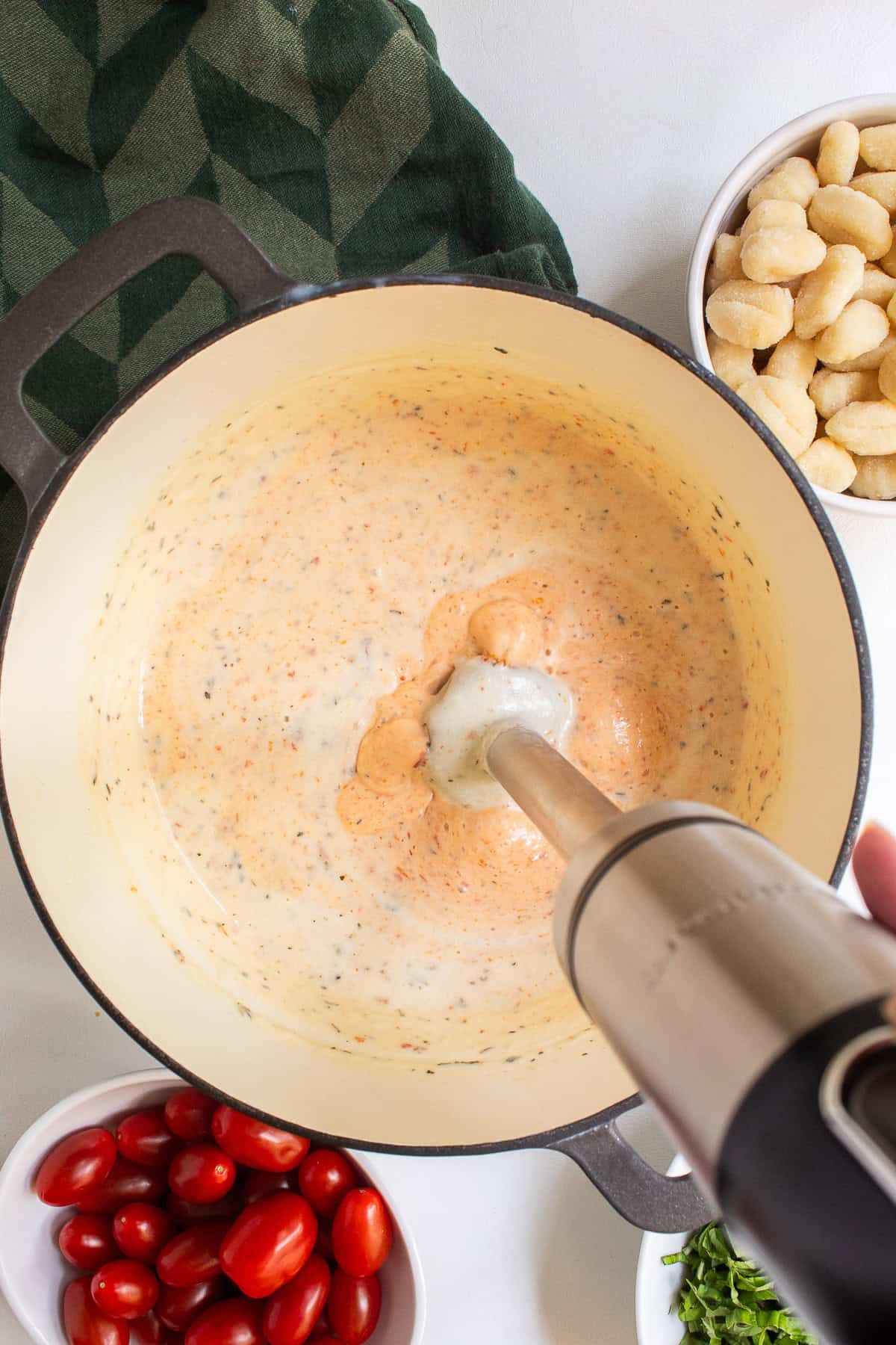 An immersion blender is blending the sauce, turning it from a cream color to a pale orange.