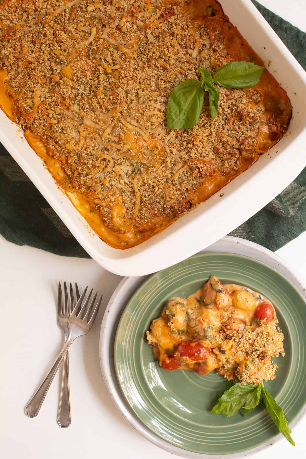 A white casserole dish with baked gnocchi is placed next to a green plate with a portion of the gnocchi.