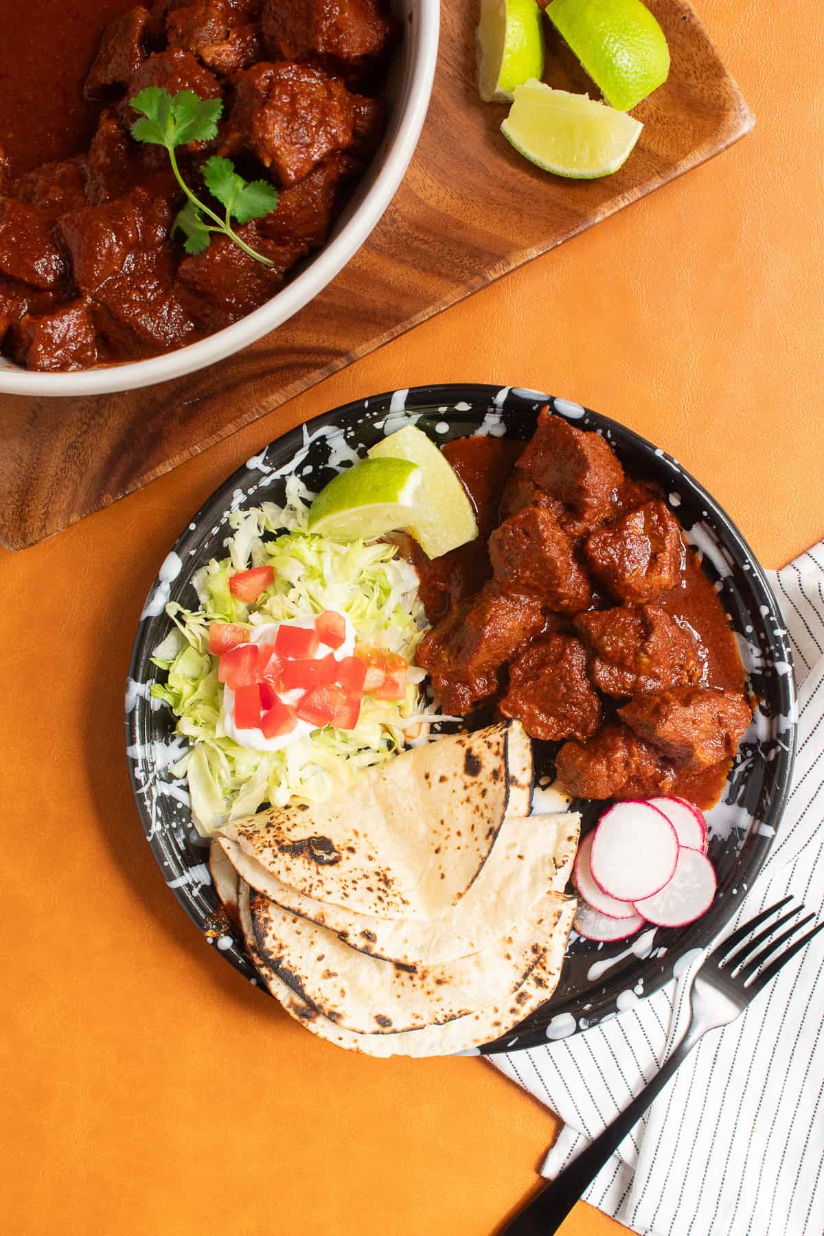 Tender pieces of pork bathed in chile colorado sauce are served with charred tortillas, radishes, limes, and shredded lettuce on a black and white plate.