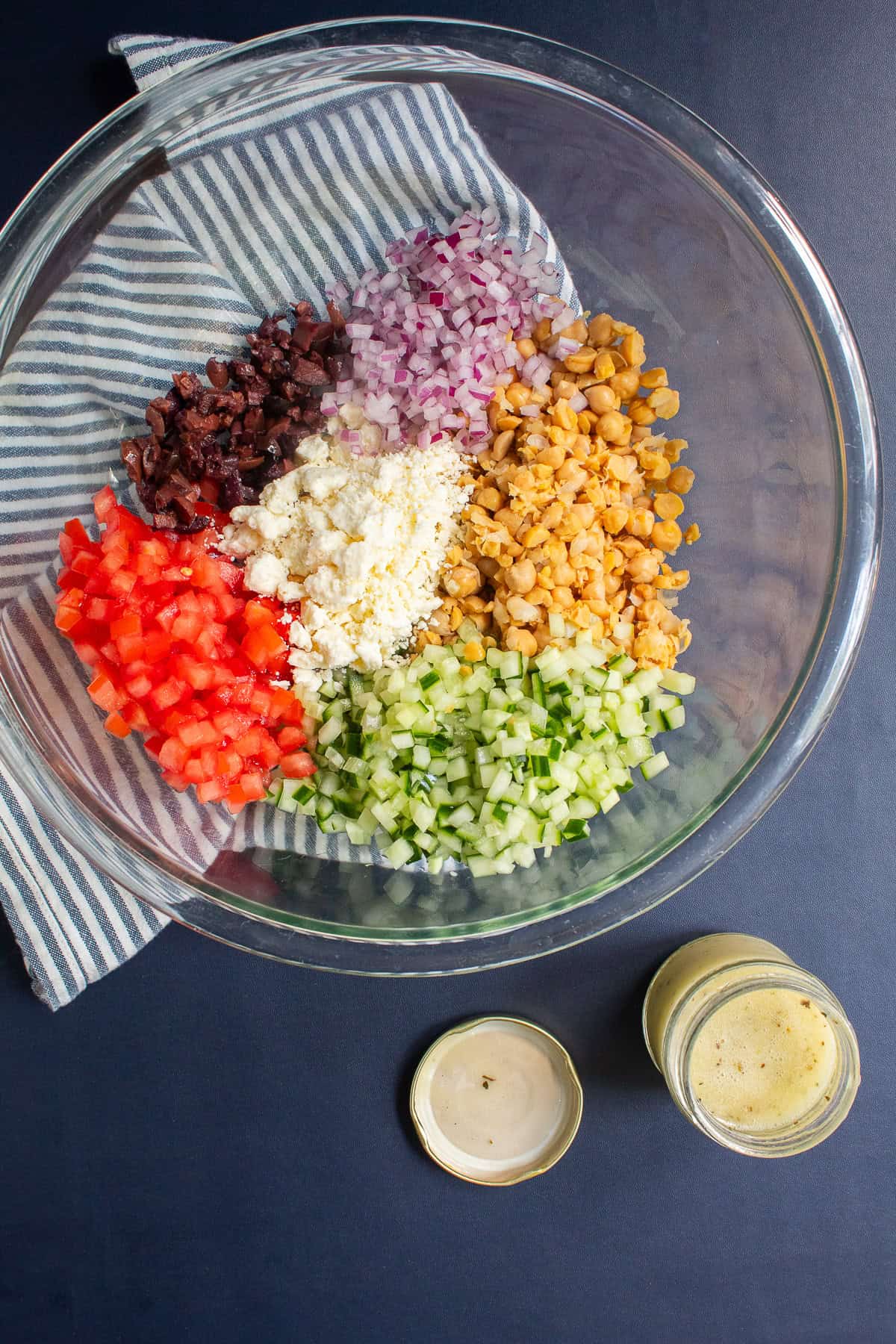 Piles of the finely diced ingredients are in a large glass bowl alongside a jar of Greek salad dressing.