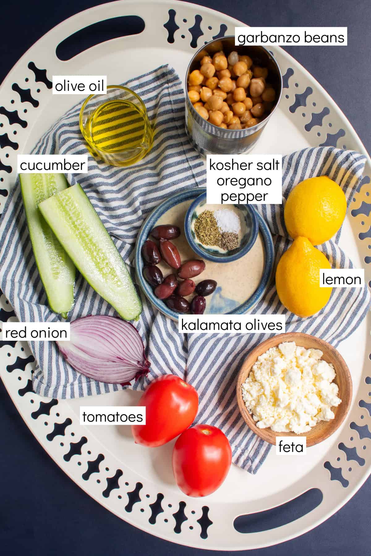 Ingredients for the recipe are displayed and labeled with white text boxes including the words olive oil, garbanzo beans, cucumber, kosher salt, oregano, pepper, lemon, red onion, kalamata olives, tomatoes, and feta.