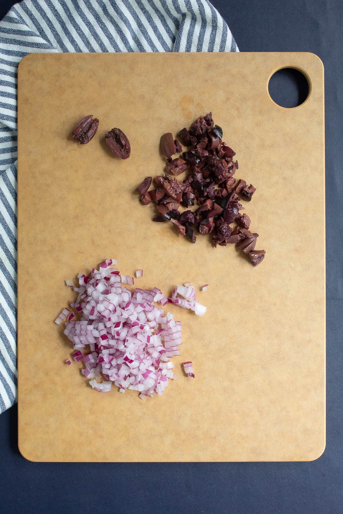 Kalamata olives are chopped next to a pile of finely diced red onion on a brown cutting board.