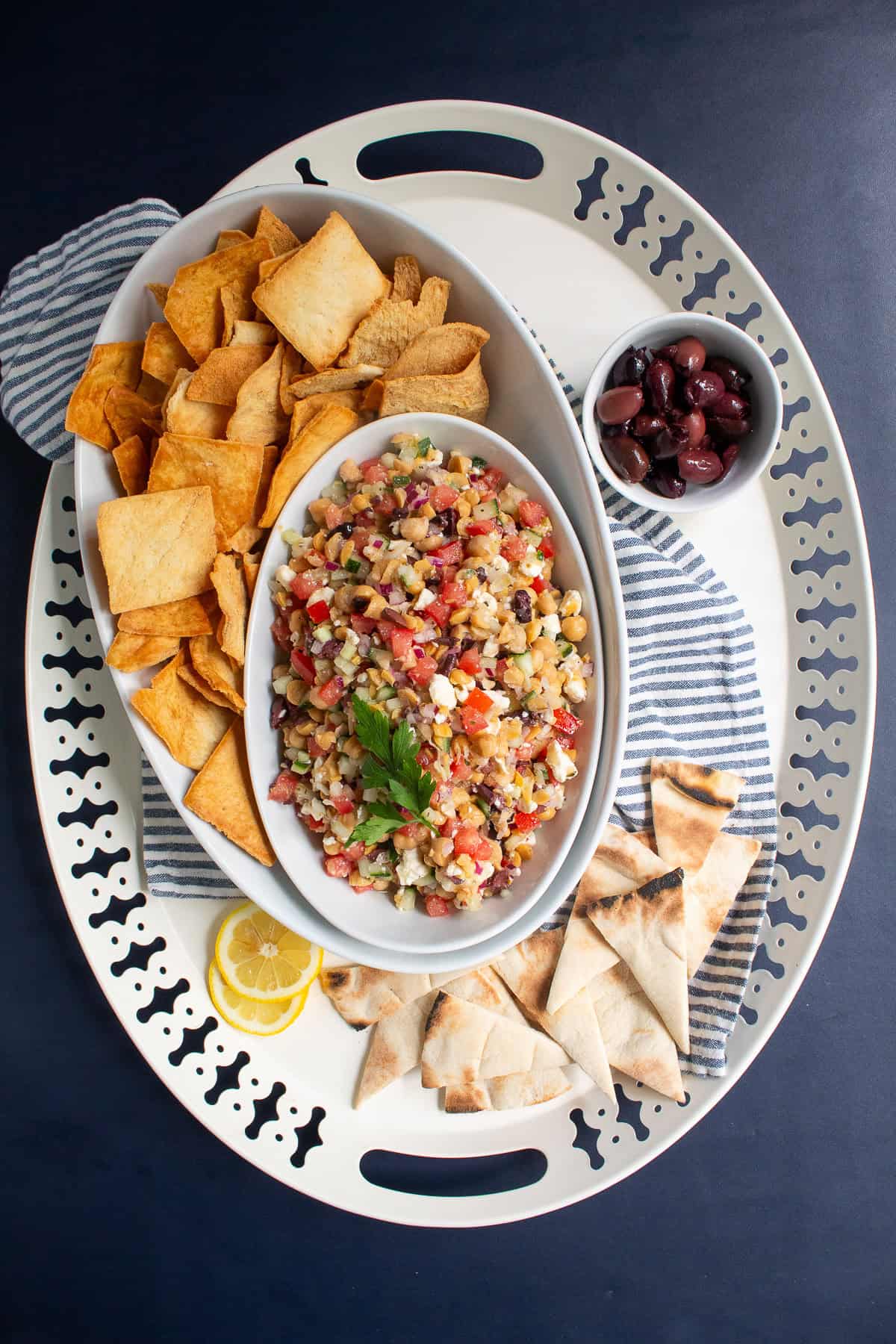The Greek salad dip is served in an oblong bowl surrounded by pita chips, lemon slices, olives, and pita triangles.